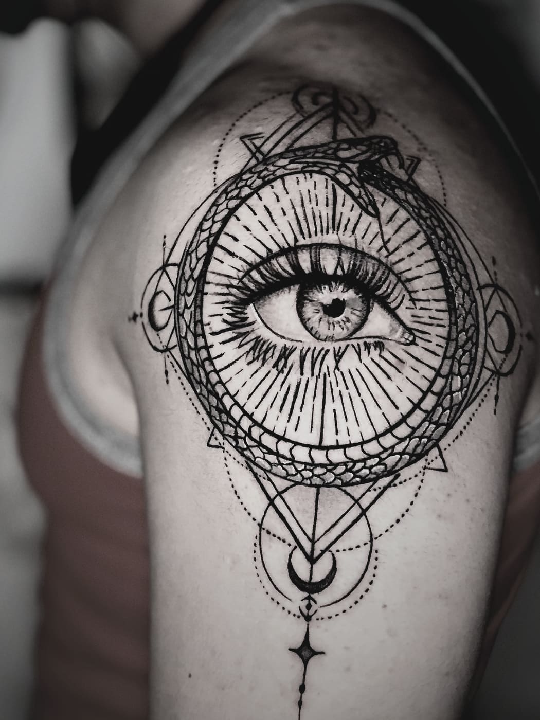 Sacred-geometry mandala on an upper arm built around a shaded realistic eye, with moons and a falling star below