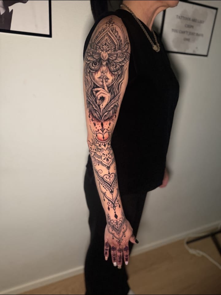 Ornamental sleeve running from shoulder to the back of the hand: a mandala at the shoulder, a moth above a face holding a finger to its lips, feathers and a heart below it, and filigree scrollwork ending in small motifs on the hand