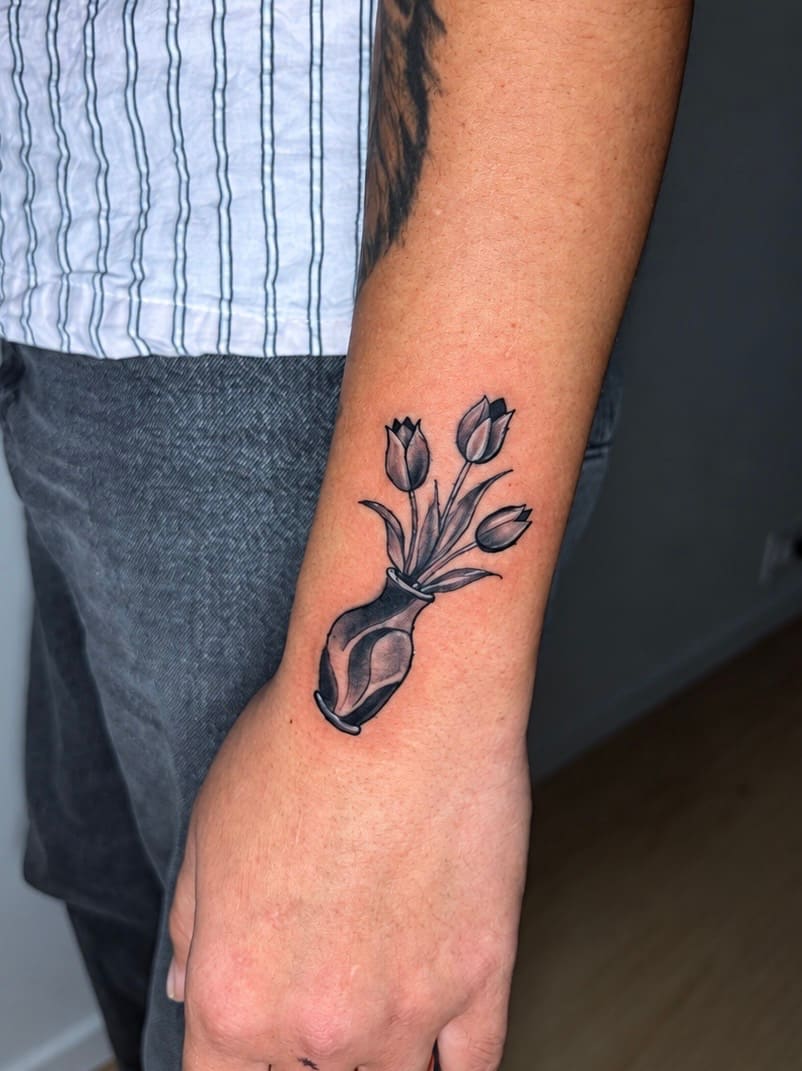 Four tulips in a shaded vase worked in black and grey down the inside of a forearm, the vase sitting at the wrist